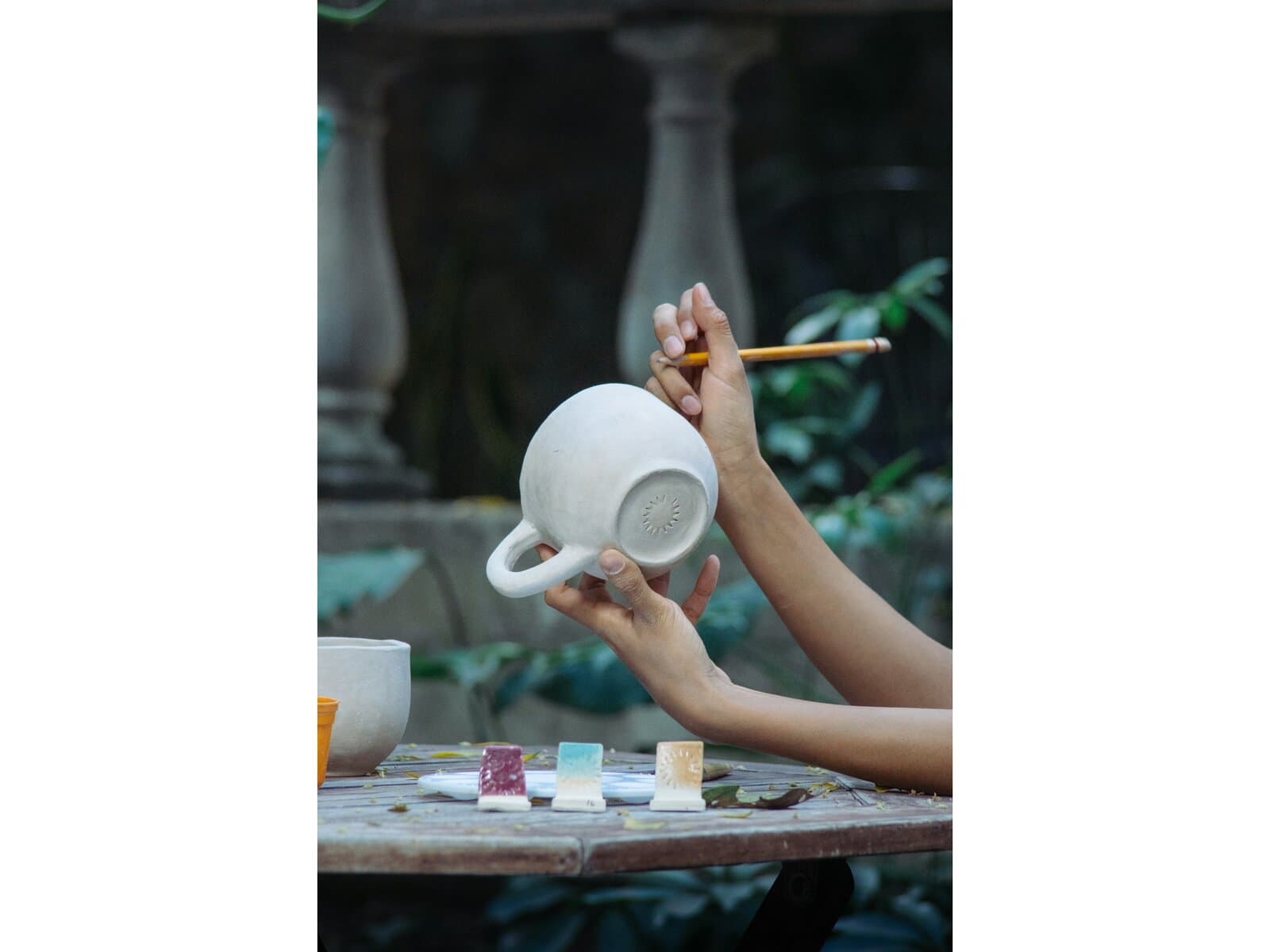 A pair of hands holds a freshly-thrown white ceramic mug above an outdoor wooden table with a paintbrush in one hand and three glaze-test tiles in front — representative of the paint-your-own-pottery sessions at Fire It! Ceramics.