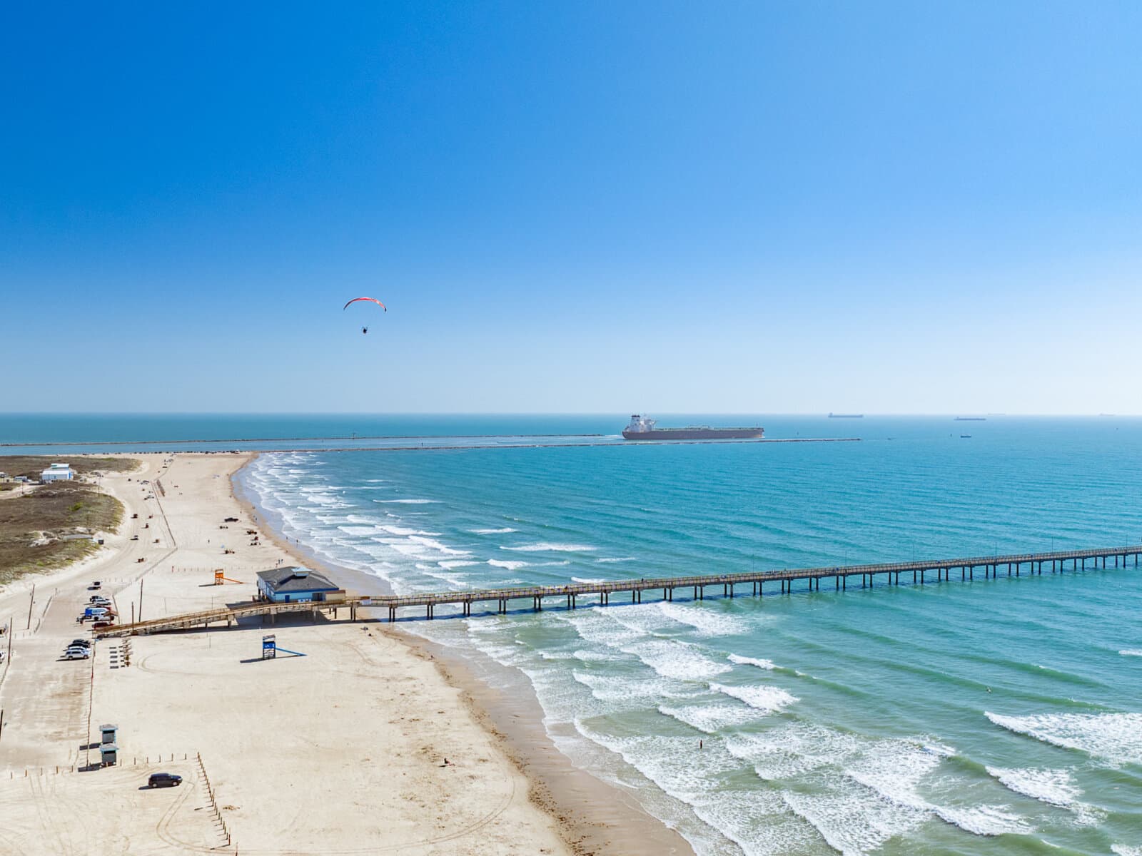 Aerial view of I.B. Magee Beach Park at Port Aransas — long fishing pier extending into the Gulf, a tanker passing the jetty channel, paraglider overhead, drive-on Gulf beach in the foreground.